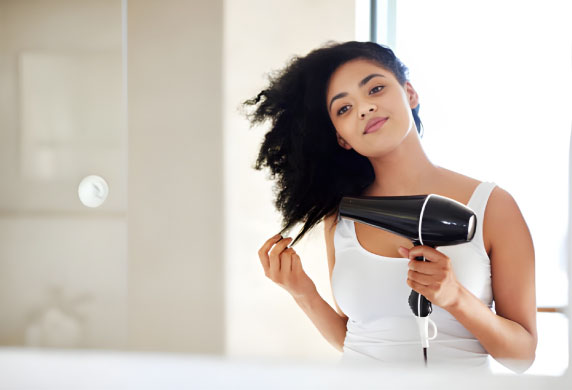 زن با موهای فر ضخیم در حال استفاده از سشوار تورمالین برای افزایش درخشندگی طبیعی م